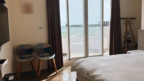Double room with white bedding, a wood headboard wall, and floor-to-ceiling windows opening onto a private balcony with lounge chairs and an open view